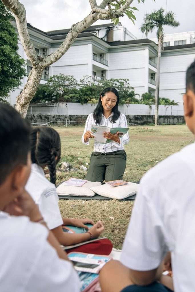 a-woman-in-white-top-holding-a-book-while-sitting-on-a-grassy-field-12719329 Teacher reading to children outdoors in a sunny setting, promoting education